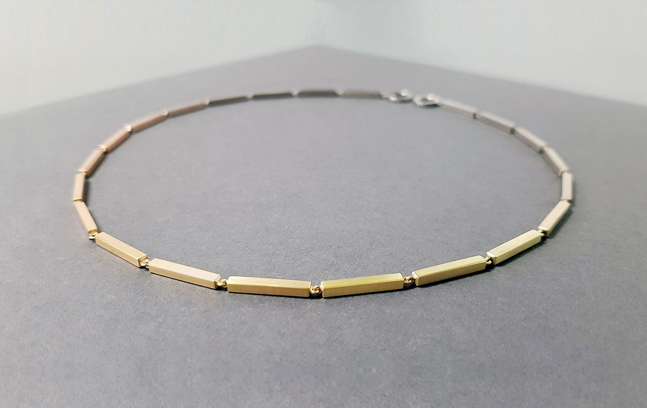 A gold necklace made of solid gold bars creating a gradiant of colour, laid on a grey background