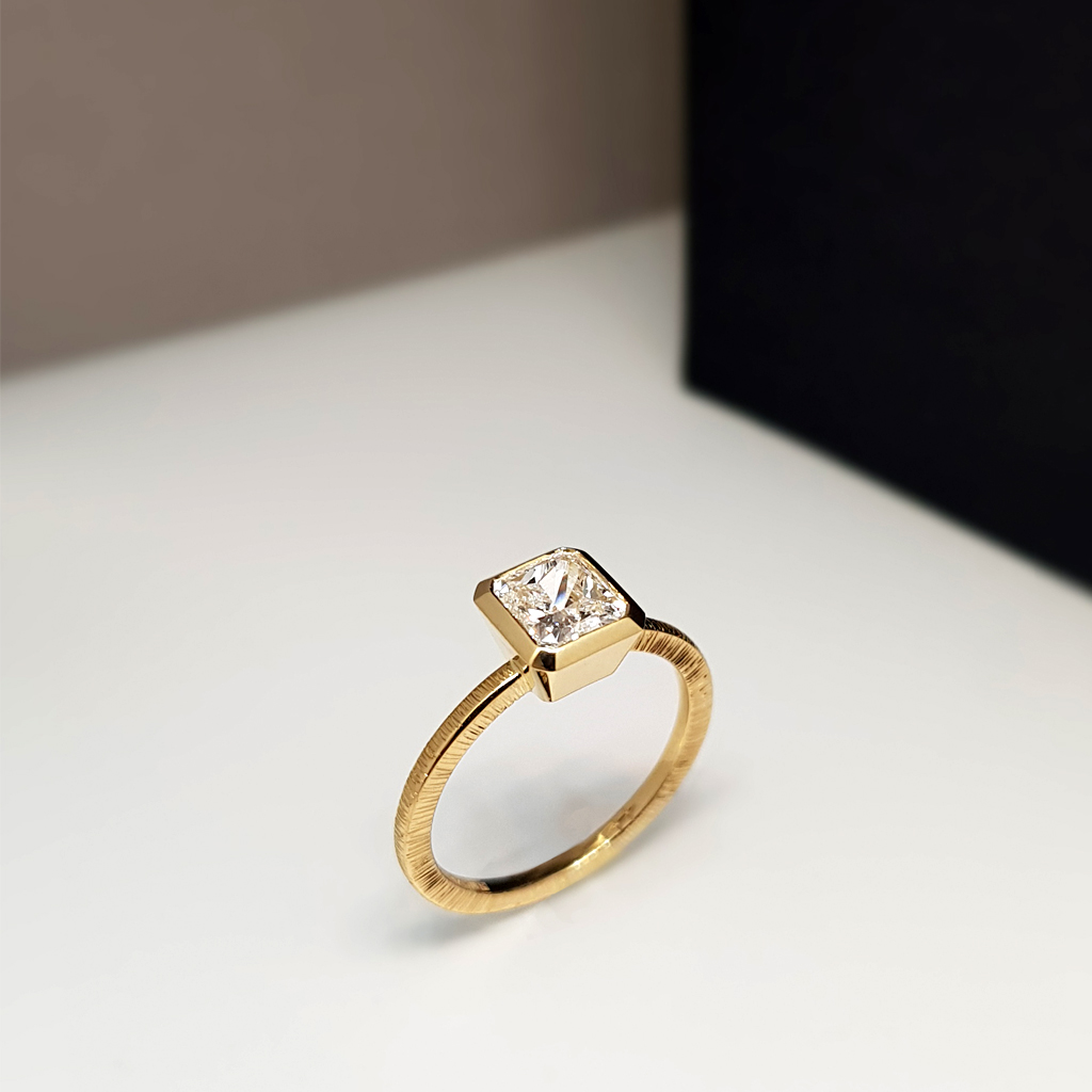 a solitaire diamond engagement ring, with a radiant cut diamond set in a yellow gold hand textured band on a white background