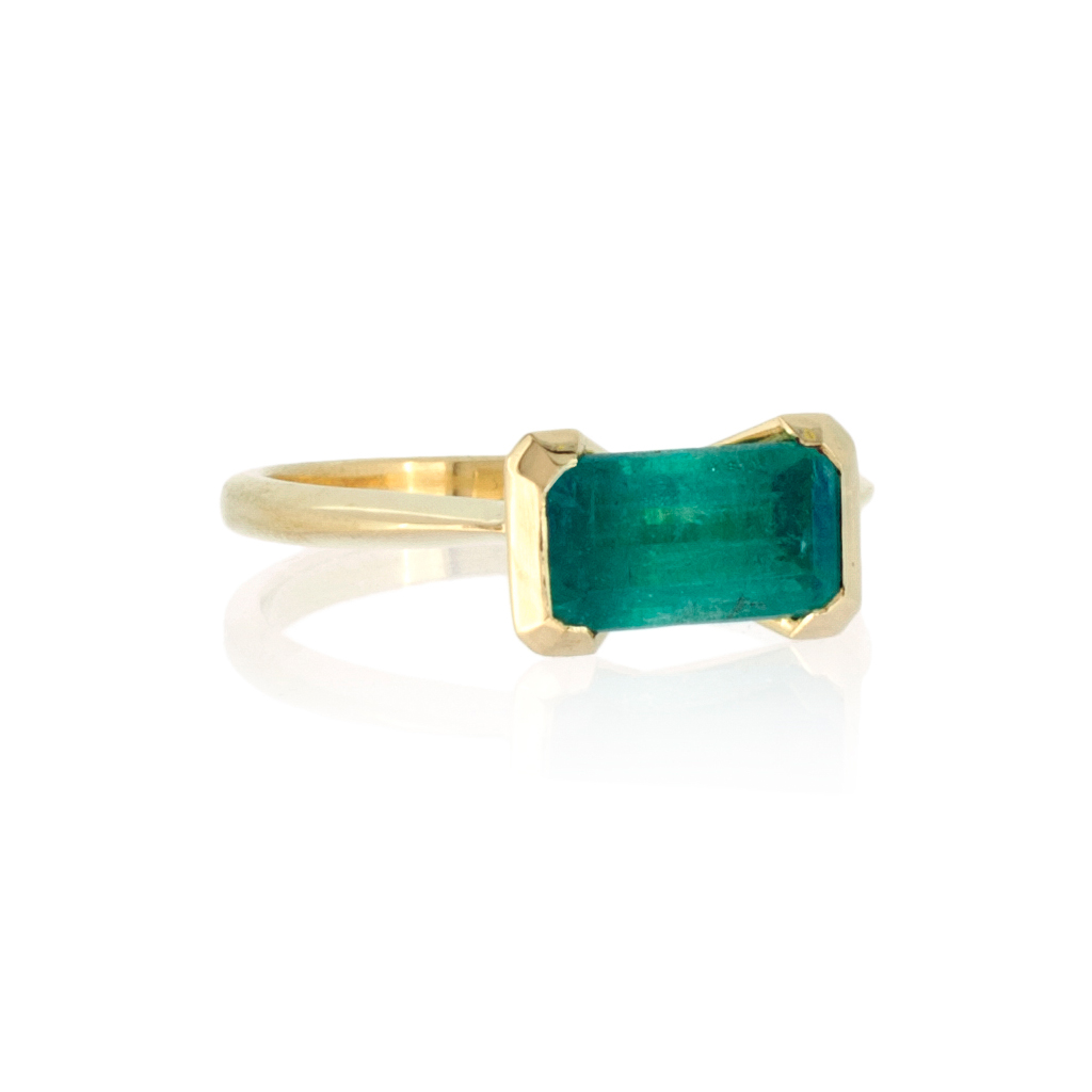 bespoke emerald art deco style engagement ring, with single rectangular emerald sent in gold on a gold band, lying on a white background