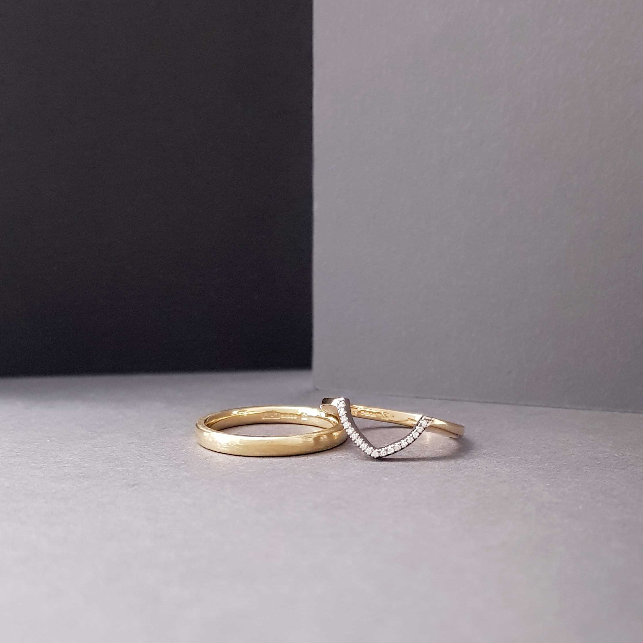 Bespoke Fairtrade Gold wishbone ring with a V-shaped detail set with diamonds resting on a man's ring. Both rings are set against a mixed backgound of grey and black with dramatic lighting.