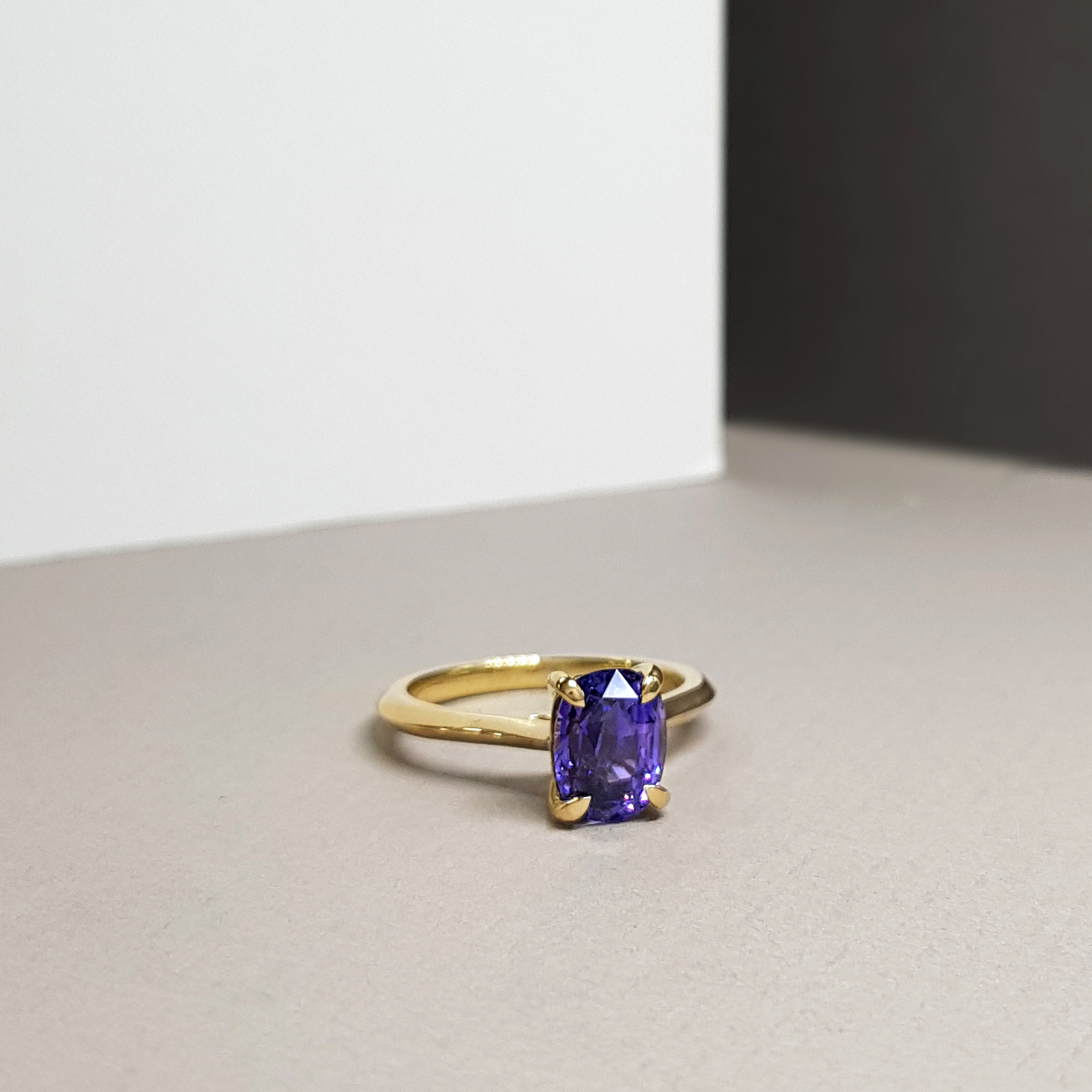 a single stone engagement ring with a large purple sapphire on a yellow fairtrade gold band, lying on a grey background