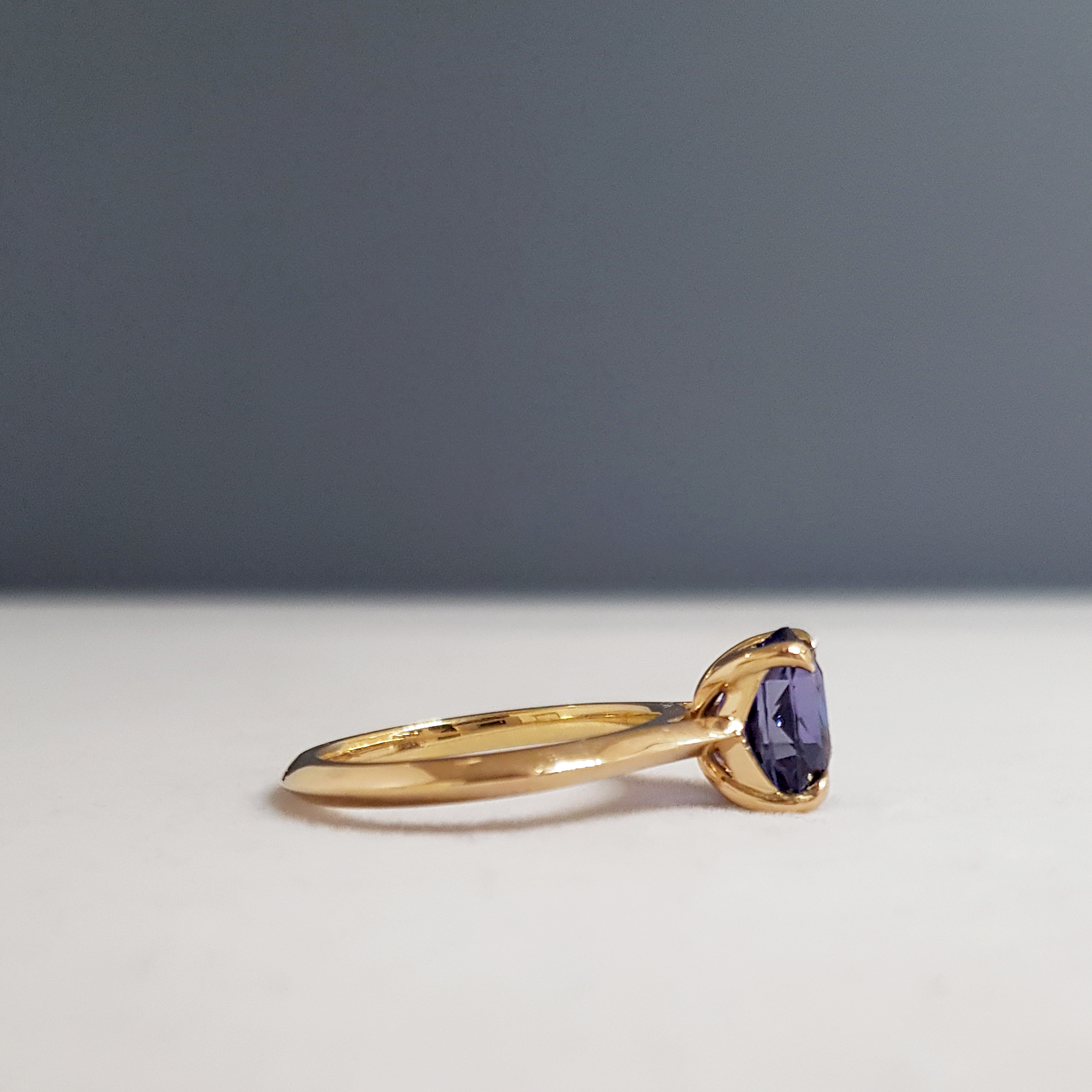 side profile of a solitaire purple sapphire egagement ring with a gold band on a grey background