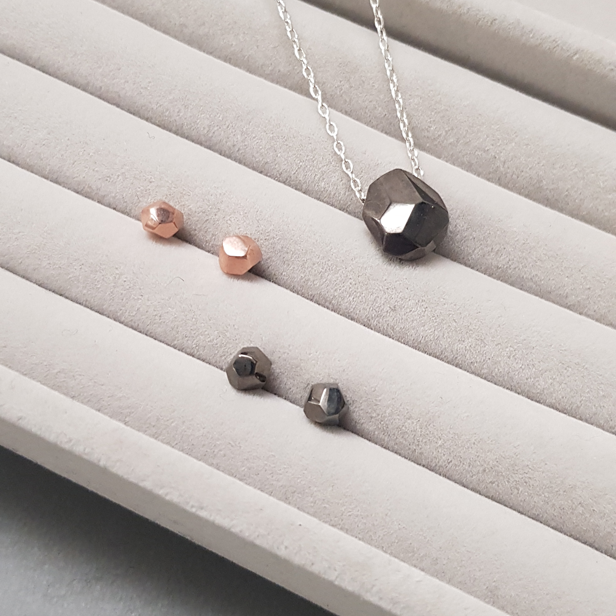two pairs of asteroid shaped earrings, one rose gold and one black, next to a polished black asteroid shaped pendant on a silver chain, on a grey fabric background