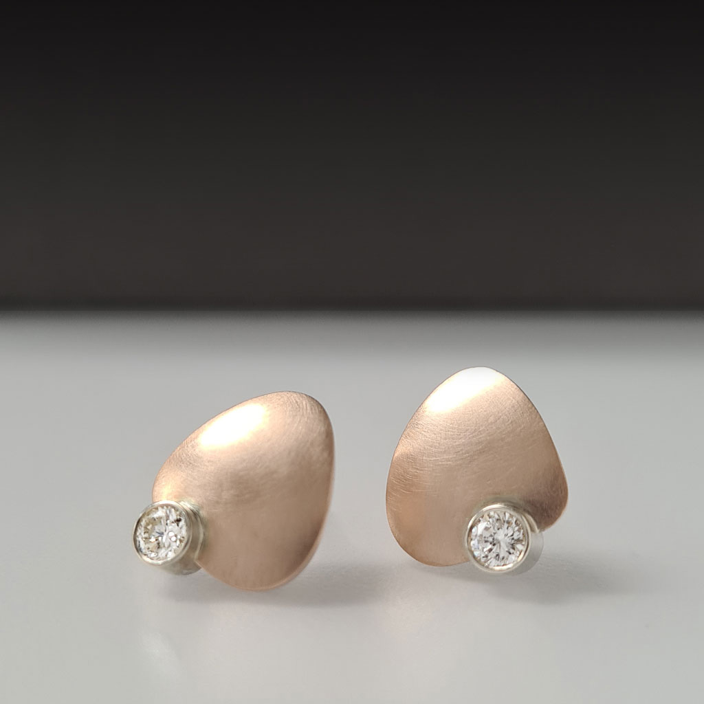 Two rose gold petal shaped earrings, each with a diamond set in white gold, sitting on a white and black background.