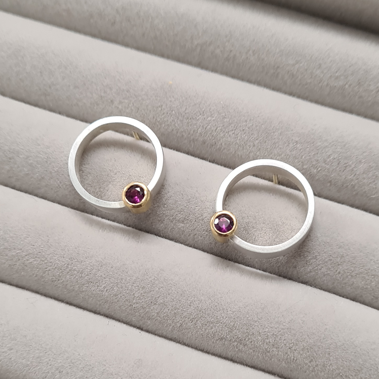 A pair of earrings with a silver band, set with a single garnet in gold, laid on a grey material jewellery presentation pad