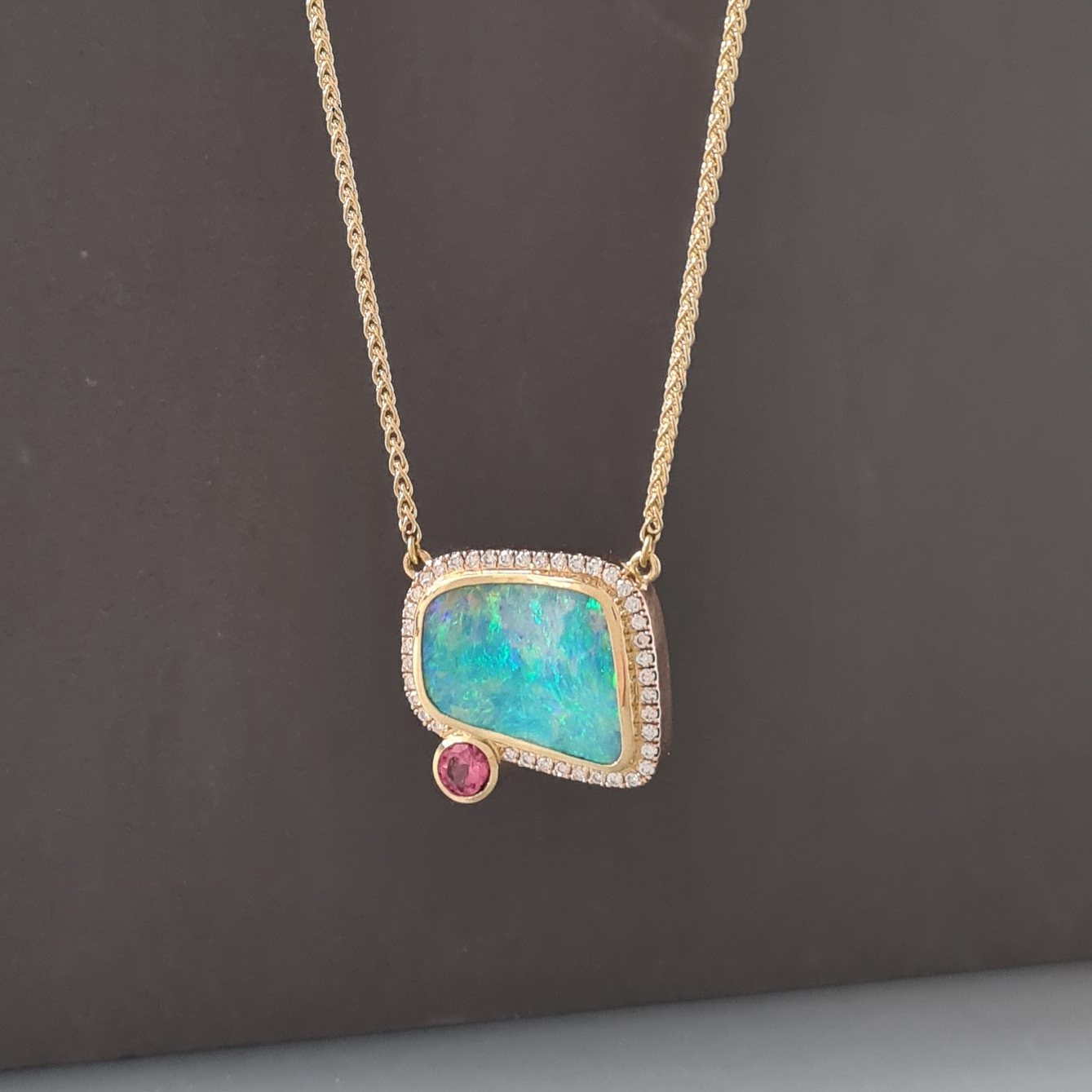 Boulder opal, diamond and padparadscha sapphire hanging on a gold chain on a dark grey background.