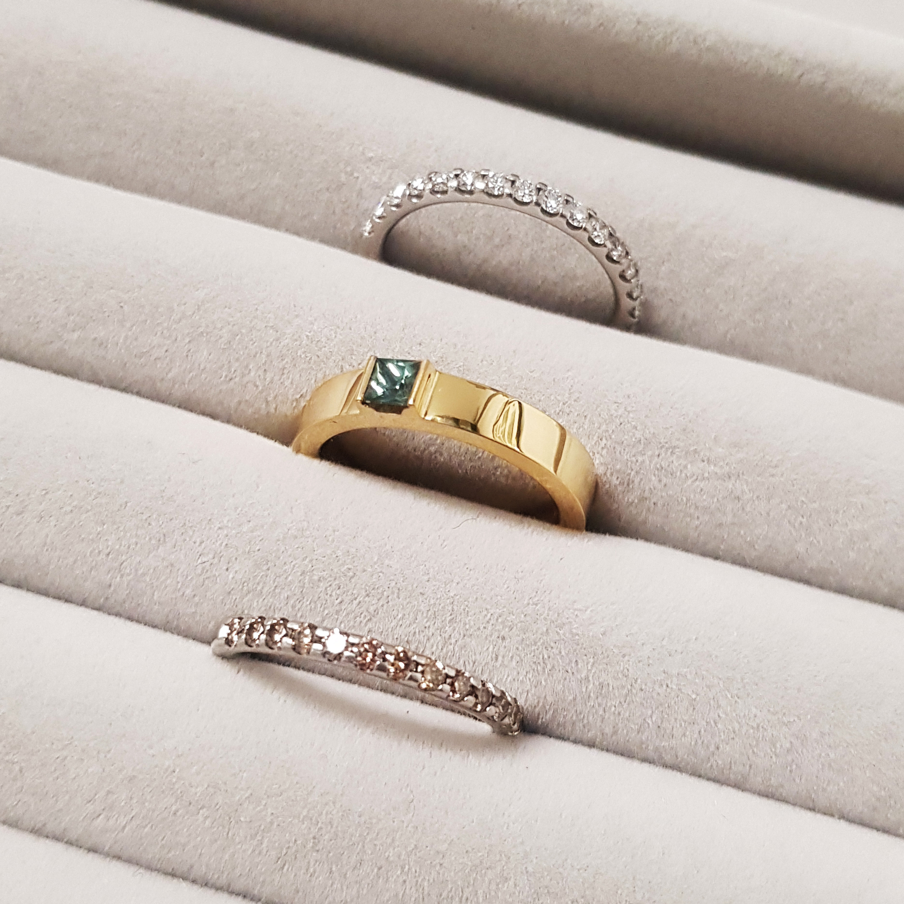 Two diamond eternity rings with an emerald and gold ring held in a row on a grey fabric display pad