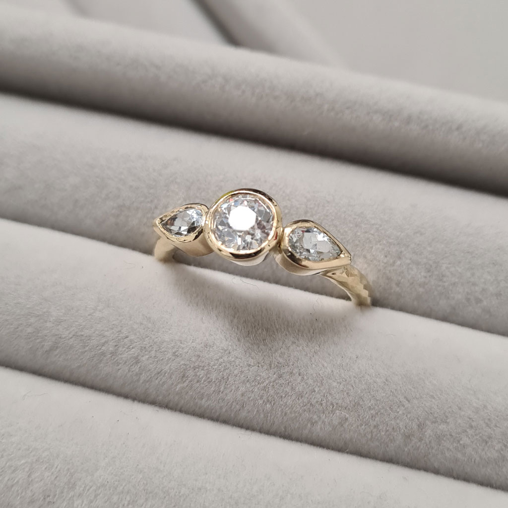 Three stone ring with round diamond and two pear cut topaz in yellow gold on a grey velvel tray.