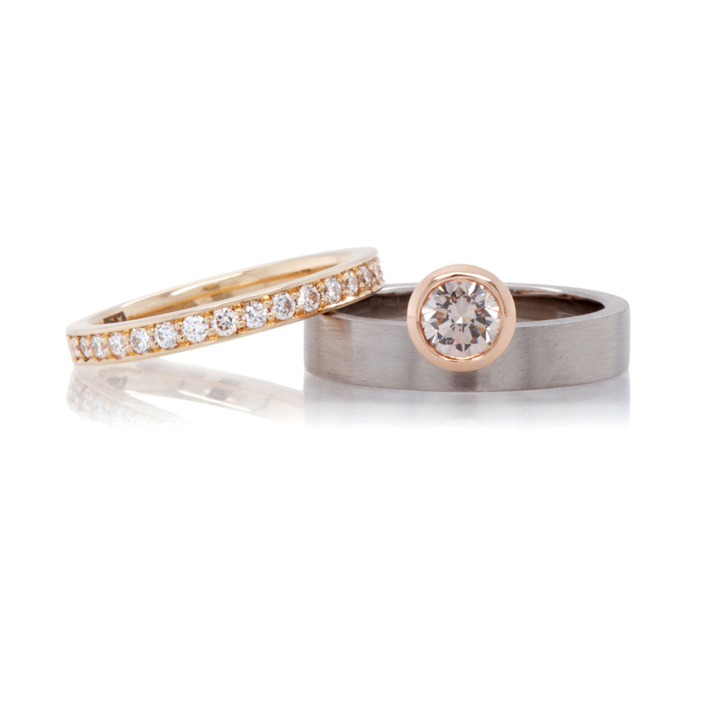 Two rings laying down on a white background with a yellow gold band resting on a diamond engagement ring.