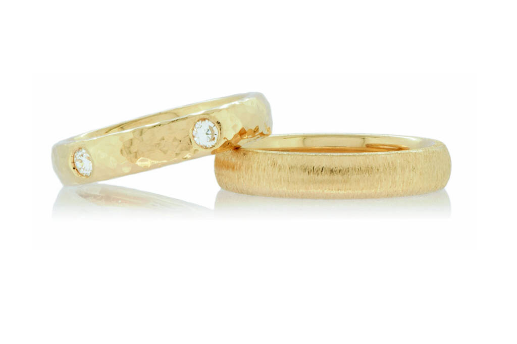 a pair of textured gold wedding bands, a gold ring with two diamonds and a hammered finish lies on top of a gold ring with a matt finish on a white background