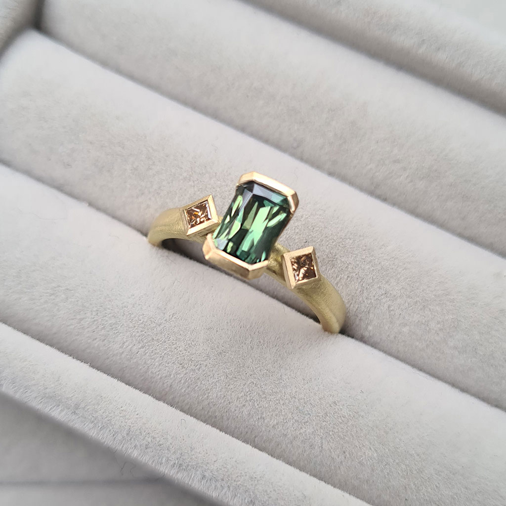 Art deco inspired engagement ring with a central green sapphire and champagne diamonds on a gold band set in a grey velvet ring tray.
