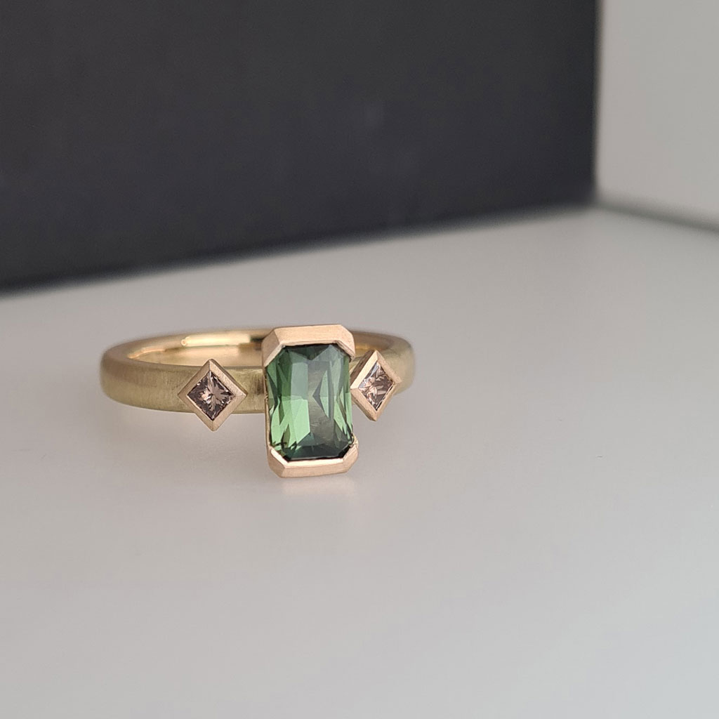 Art deco inspired engagement ring with a central green sapphire and champagne diamonds on a gold band against a black and white colour block background