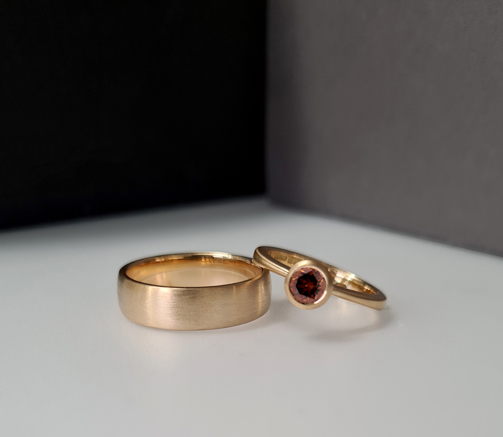 Two blush gold rings on a colour block background of white, grey and black. The ring on the right is a diamond solitaire ring resting on a 6mm wide wedding band to the left. Both rings have a satin matte finish.