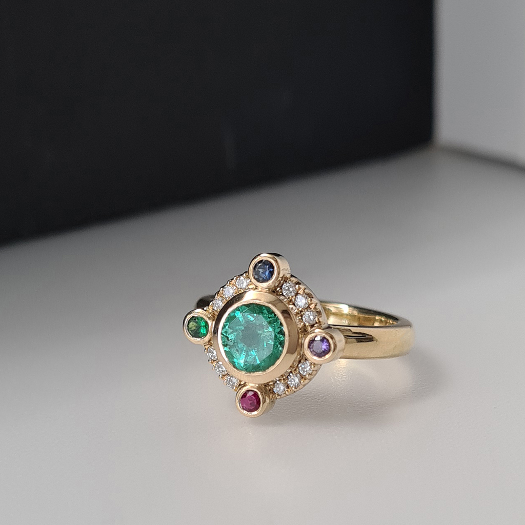 A round green-blue Paraiba tourmaline with 4 coloured gemstones set at the compass points around the around the tourmaline and small diamonds in between. The ring is made in 18k yellow gold and placed on a black and white colour block background..