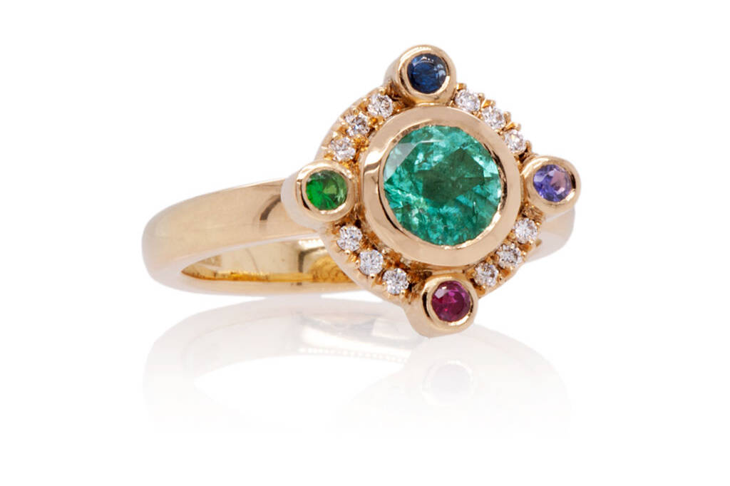 A round green-blue Paraiba tourmaline with 4 coloured gemstones set at the compass points around the around the tourmaline and small diamonds in between. The ring is made in 18k yellow gold and placed on a white background.