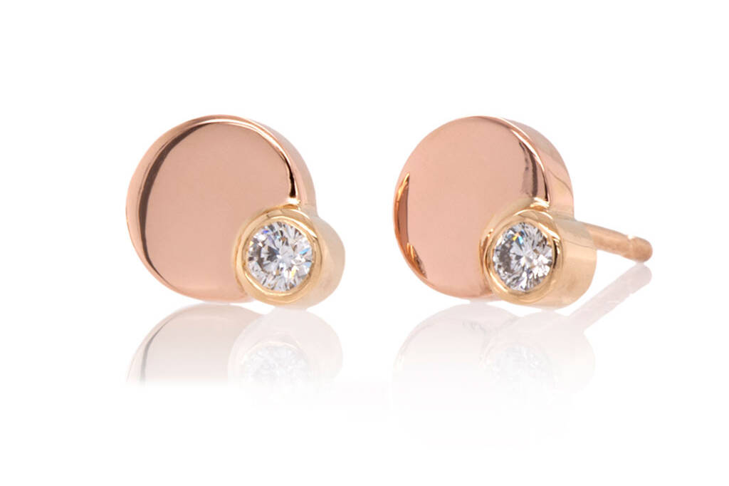 A pair of circular gold stud earrings in rose and yellow gold each set with a single diamond on a white surface and a black background.