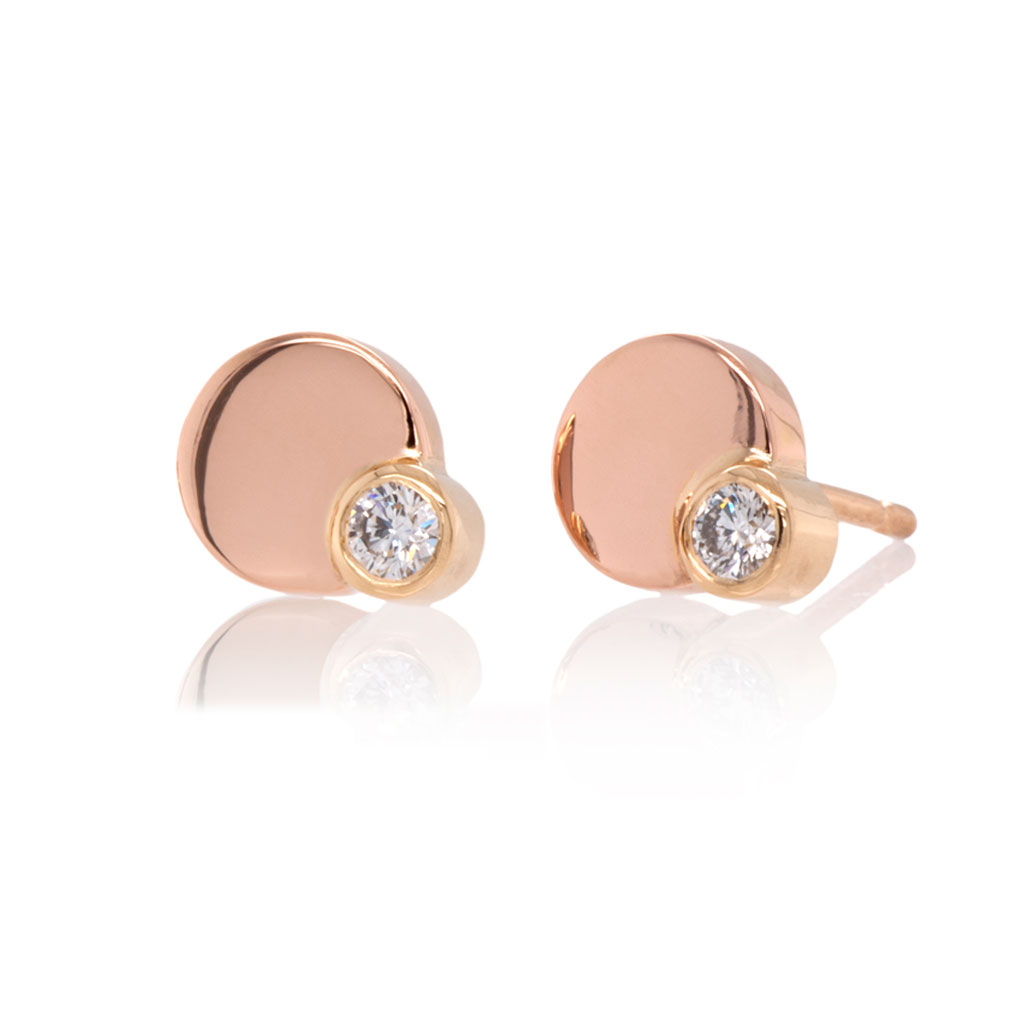 A pair of circular gold stud earrings in rose and yellow gold each set with a single diamond on a white surface and a black background.