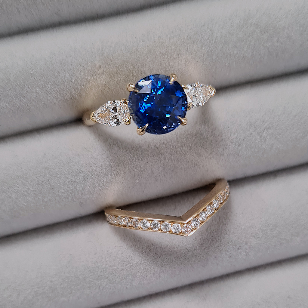 Three stone engagement ring with a blue sapphire and pear cut diamonds with a matching diamond wedding band on a grey velvet tray.