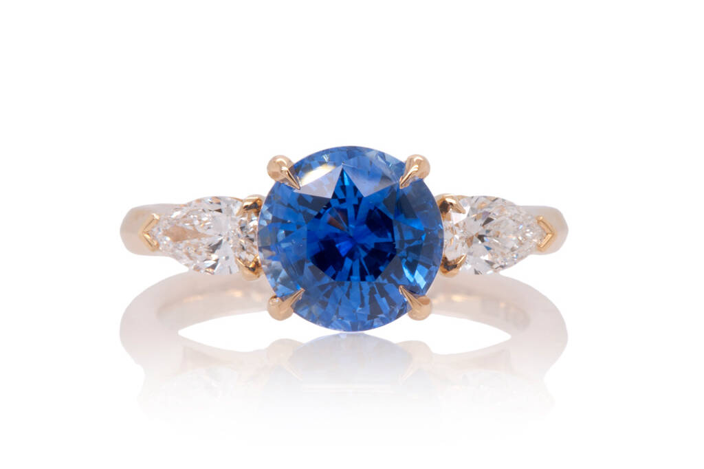 A three stone engagement ring with a central round, blue sapphire with two pear cut diamonds either side set in 18k yellow gold on a white background.