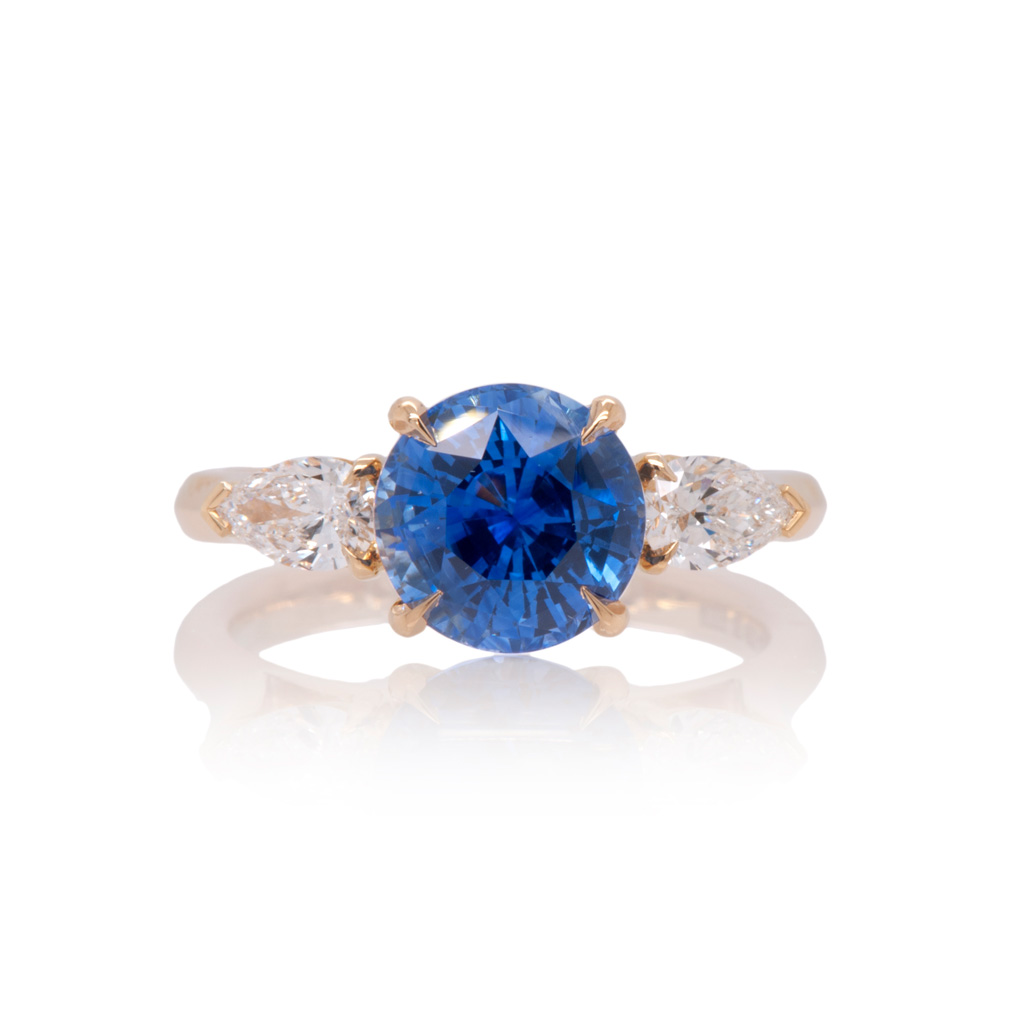 A three stone engagement ring with a central round, blue sapphire with two pear cut diamonds either side set in 18k yellow gold on a white background.