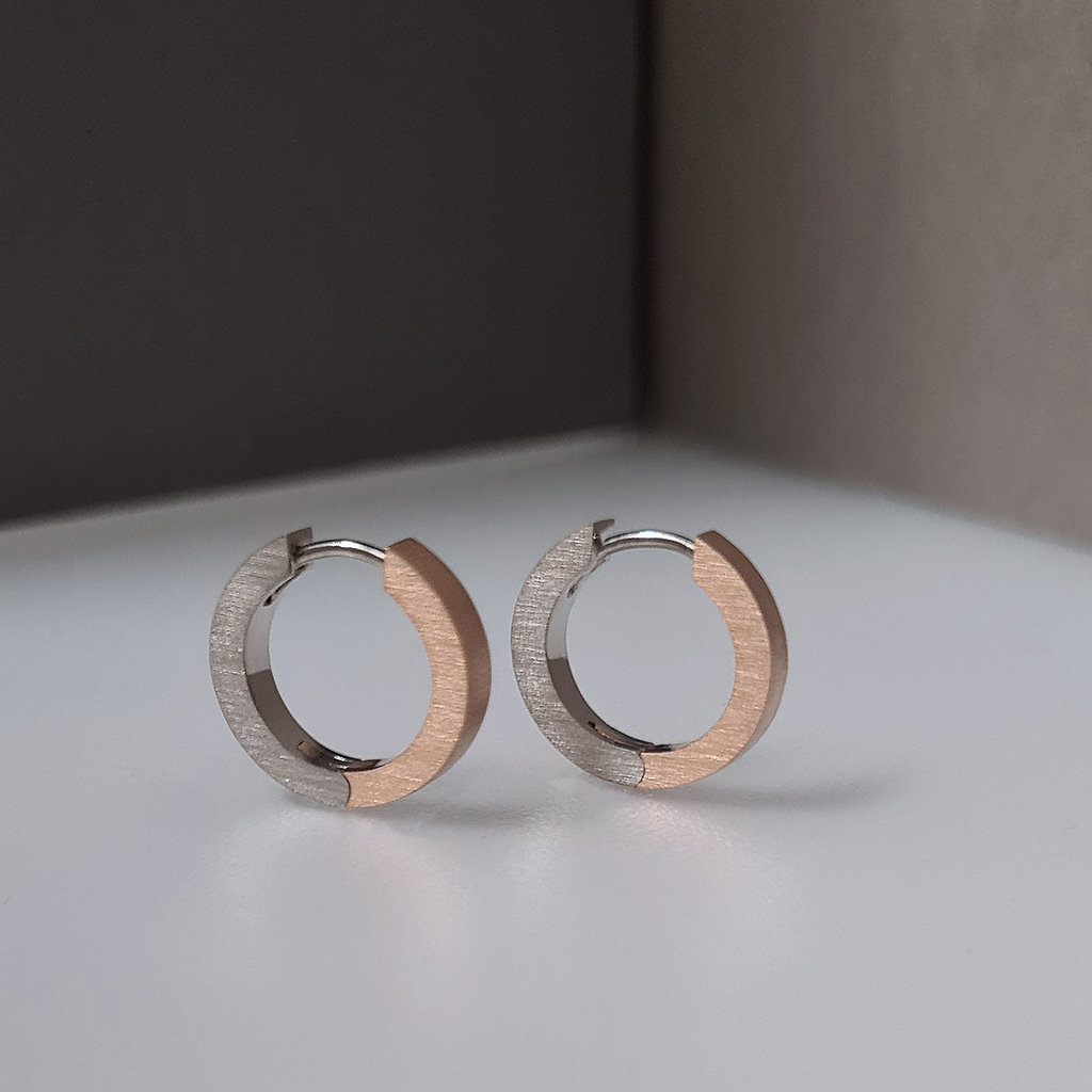 Side view of a pair of two tone hinged hoop earrings standing upright against a colour black background of black, white and grey.
