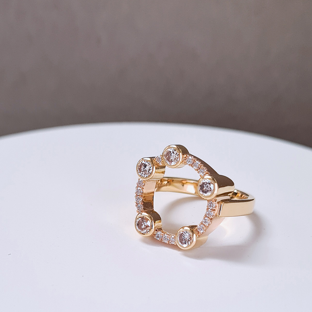 A ring with a circle of diamonds set in yellow and red gold sitting on a white plinth with a grey background.