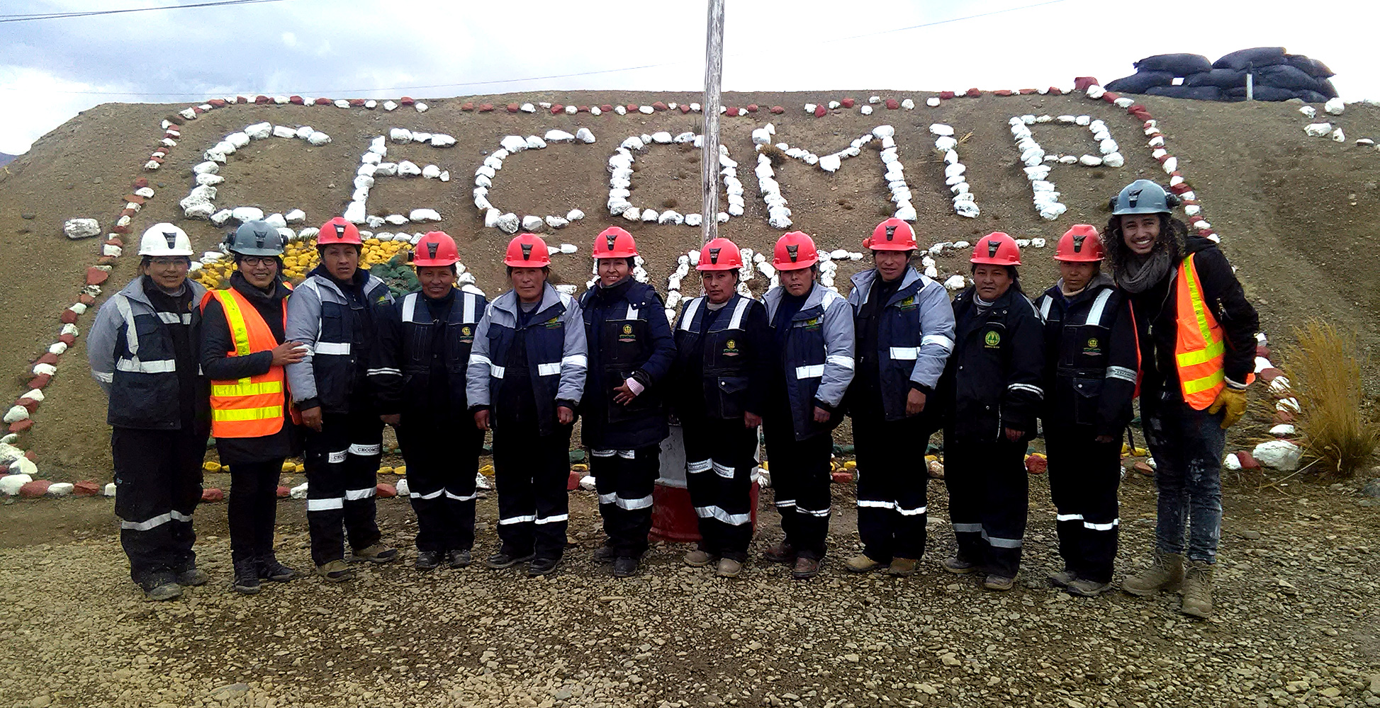12 miners in safety uniforms standing in front of a hillside with a sign made of rocks spelling out 'CiECOMIP,'