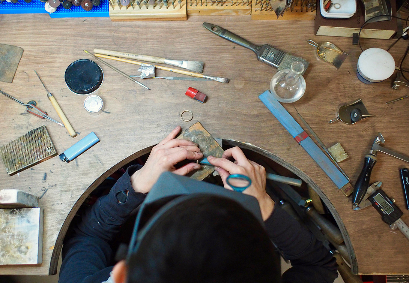 Birdseye view of Amanda's head and hands working at her jewellery bench with tools and material spread over the surface.