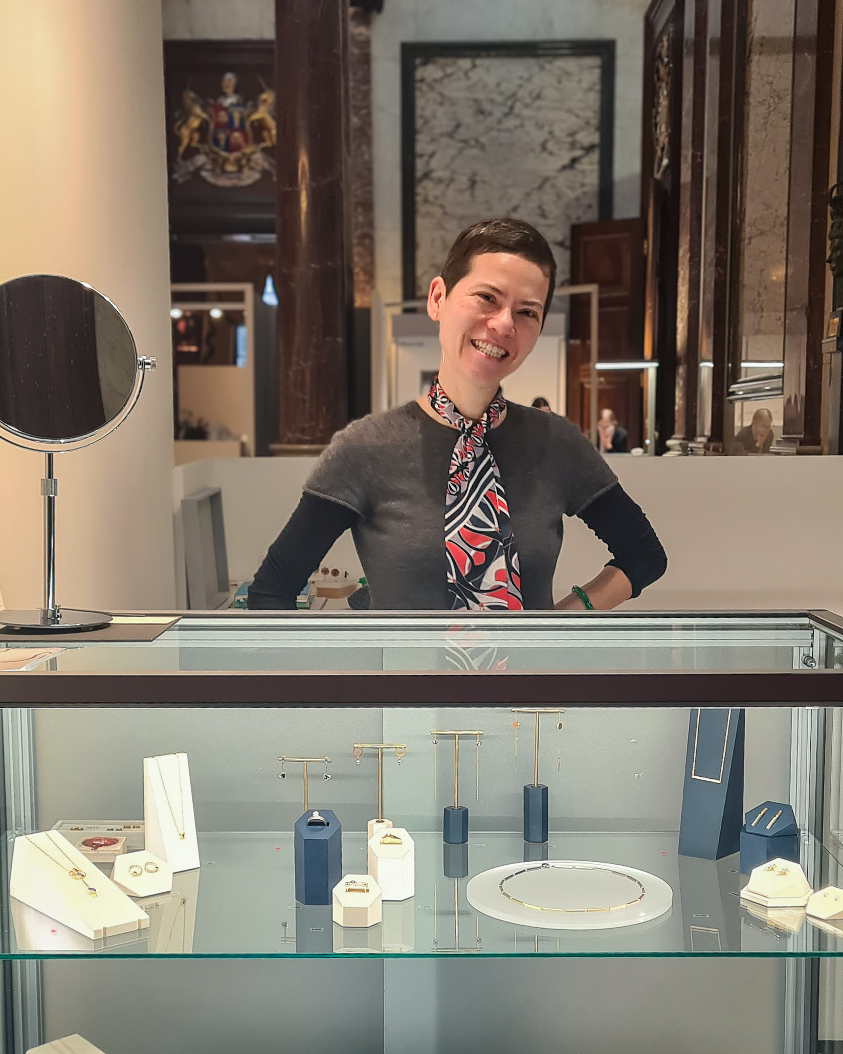 Amanda standing behind her glass display cabinet filled with her jewellery. Behind her is the open space of Goldsmiths' Hall atrium over the grand staircase